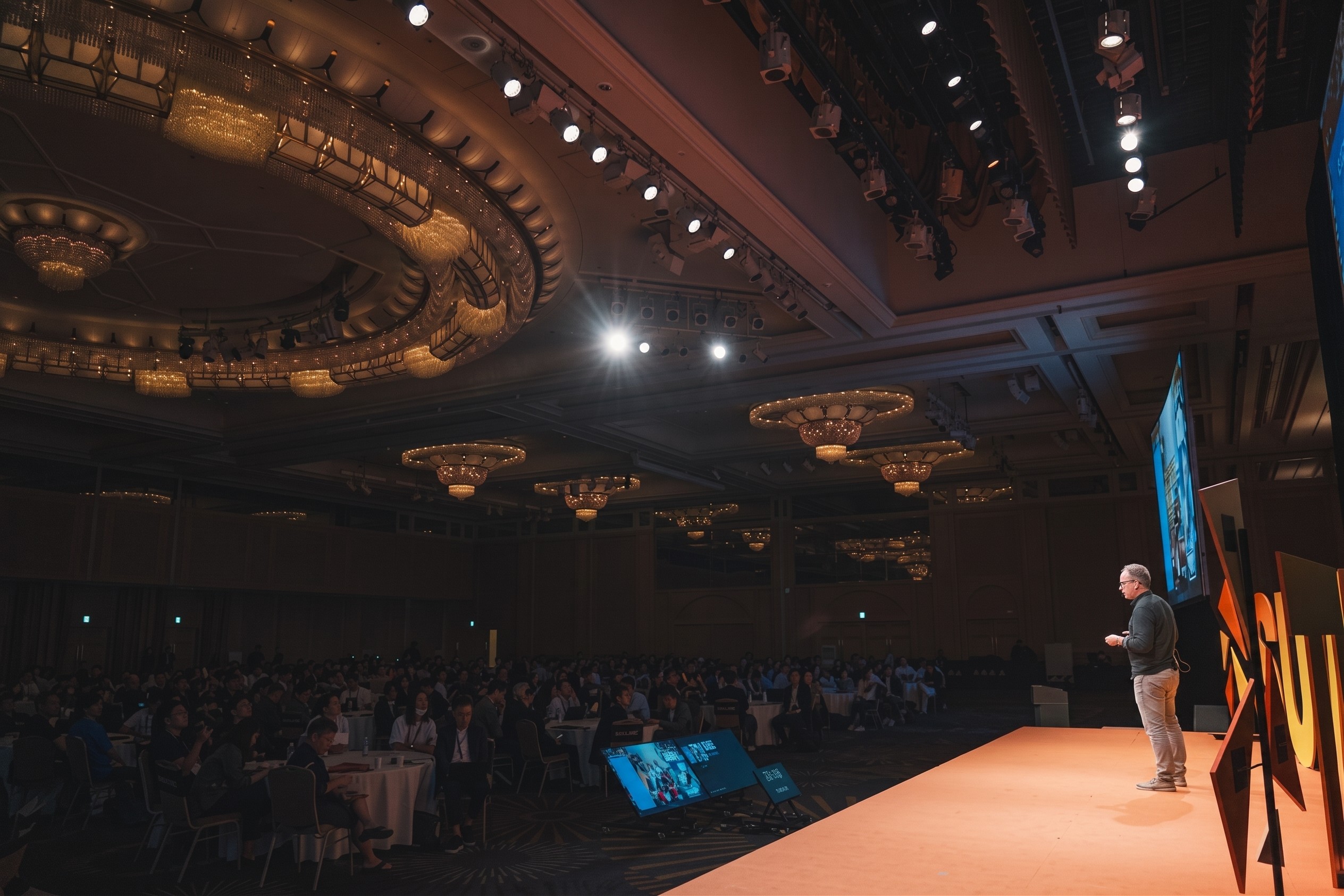 A speaker on stage in the summit ballroom, the audience seated at round tables below.